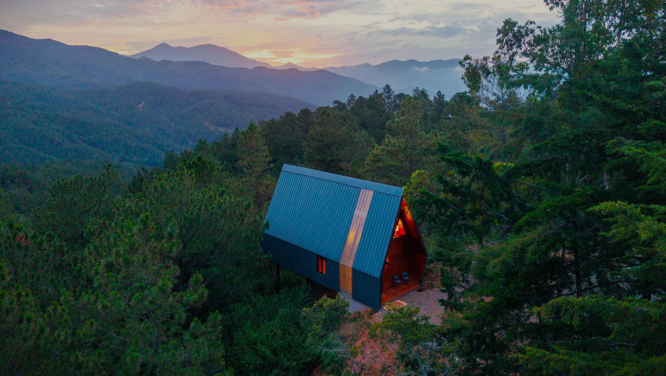 Aerial view at dusk of an Estancia Natura A-Frame cabin among pine forest, with the Cordillera Central mountains beyond